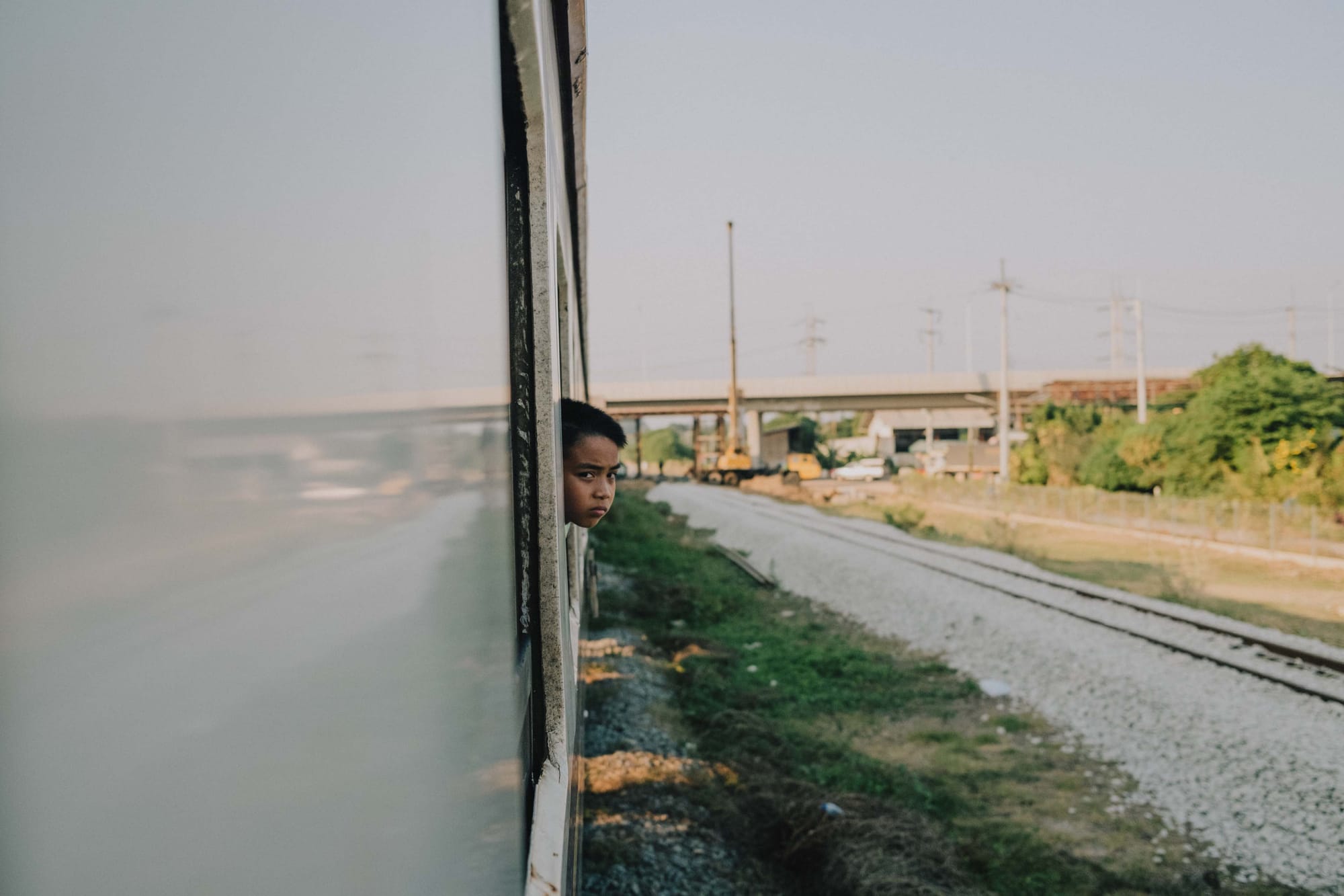A child sticks his head out of the window of a moving train in Thailand with the rural landscape in the background
